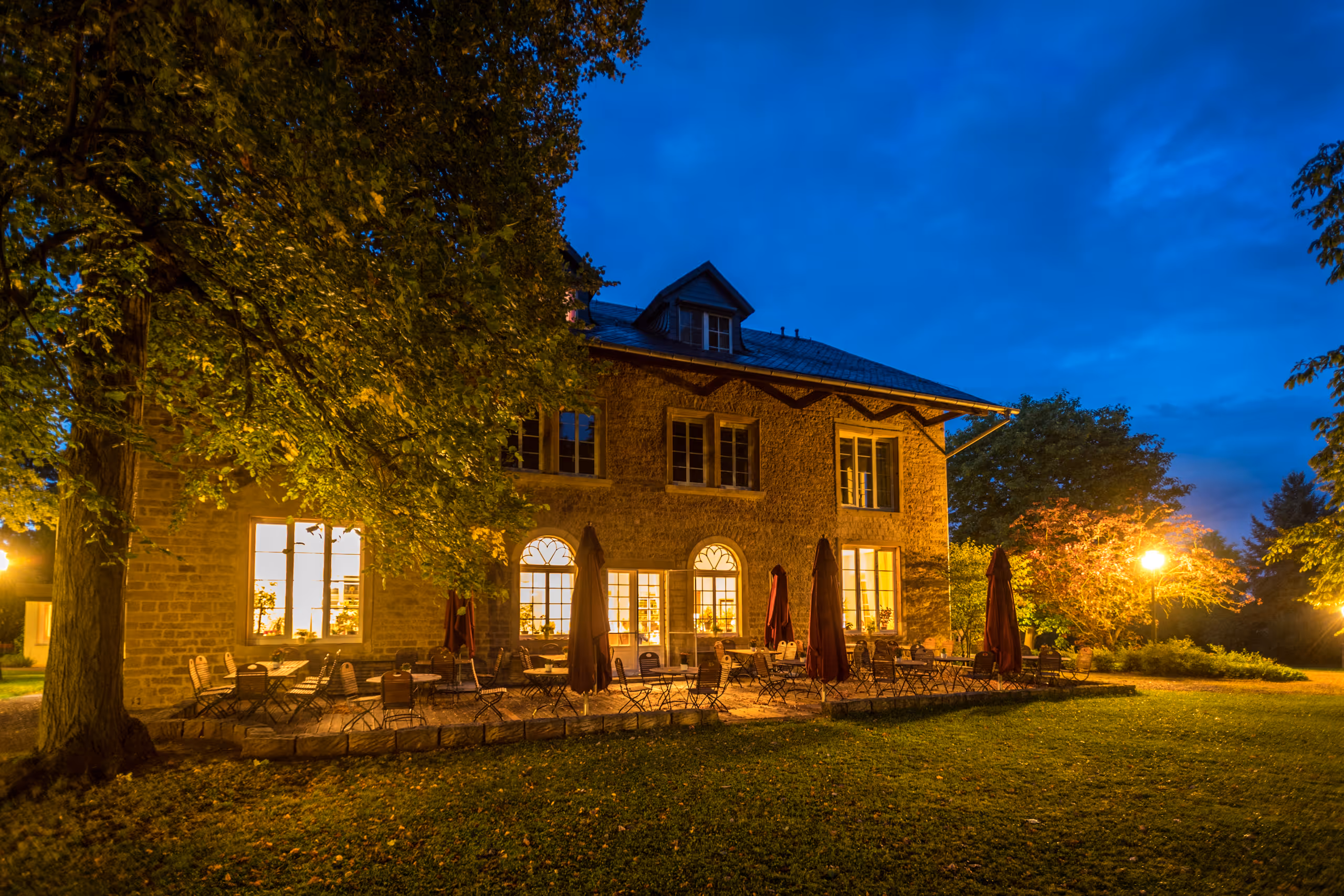 Rustic stone building with glowing windows and outdoor seating at twilight