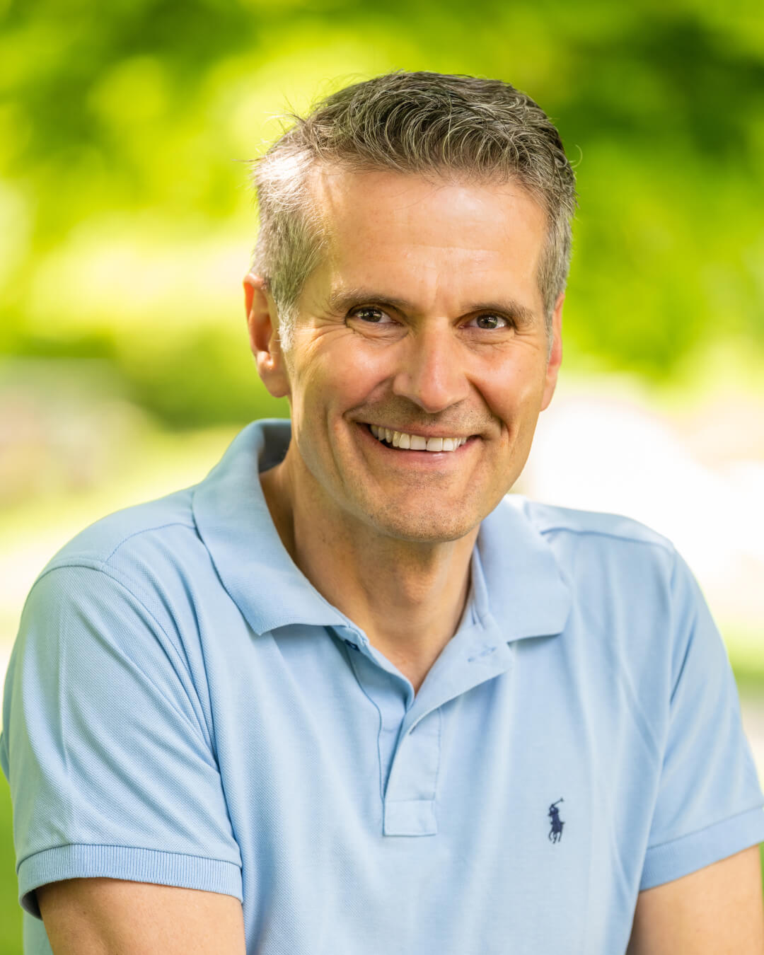 Middle-aged man in light blue polo smiling outdoors with green background