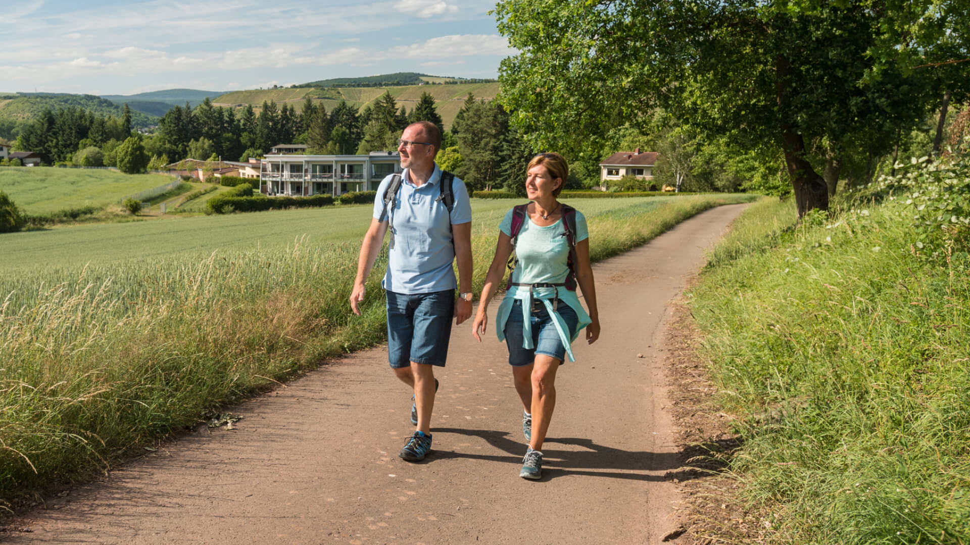 Two hikers walking on a country path surrounded by green fields and trees