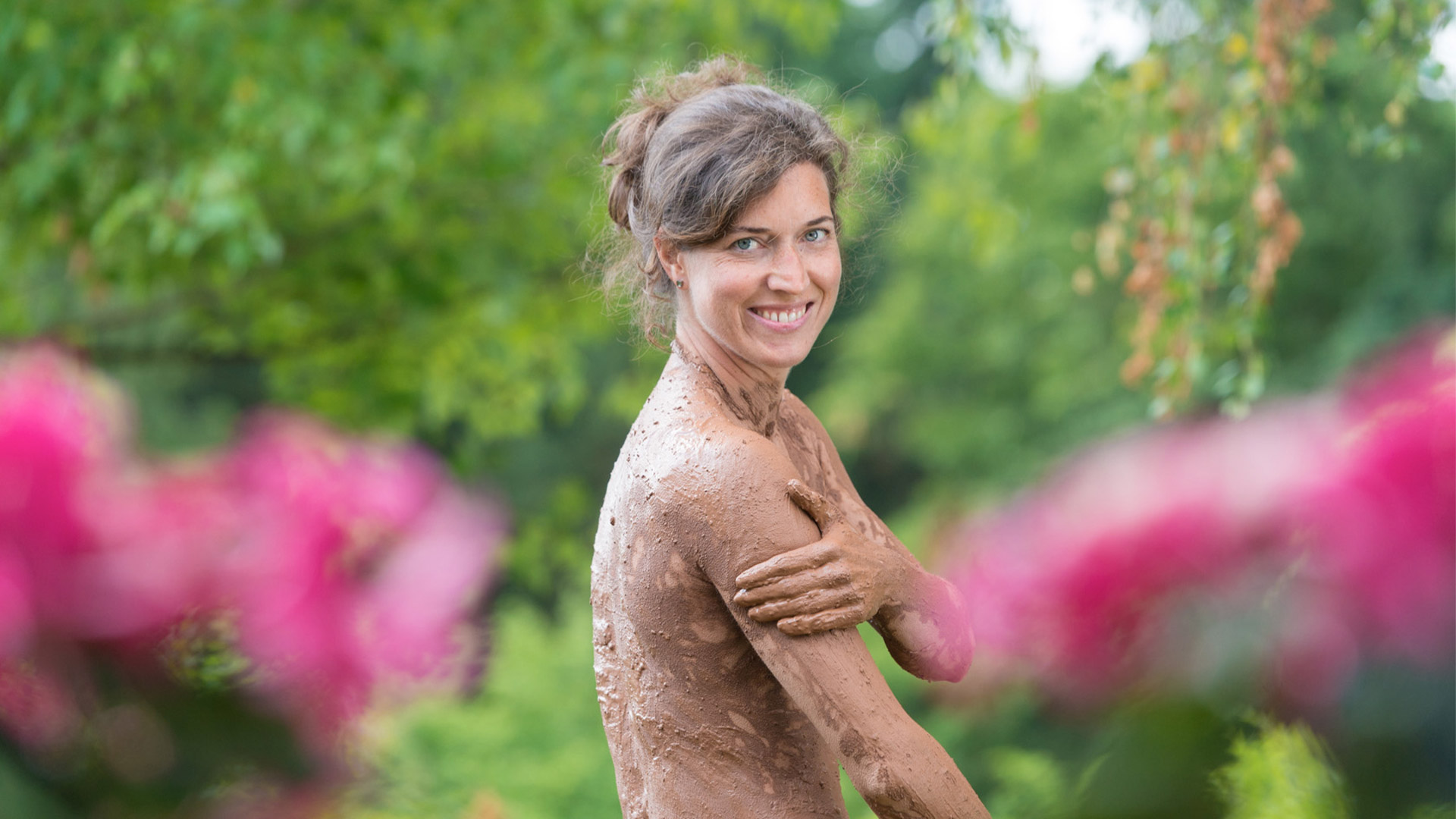 Lächelnde Person mit Lehm bedeckt, umgeben von grüner Natur und Blumen