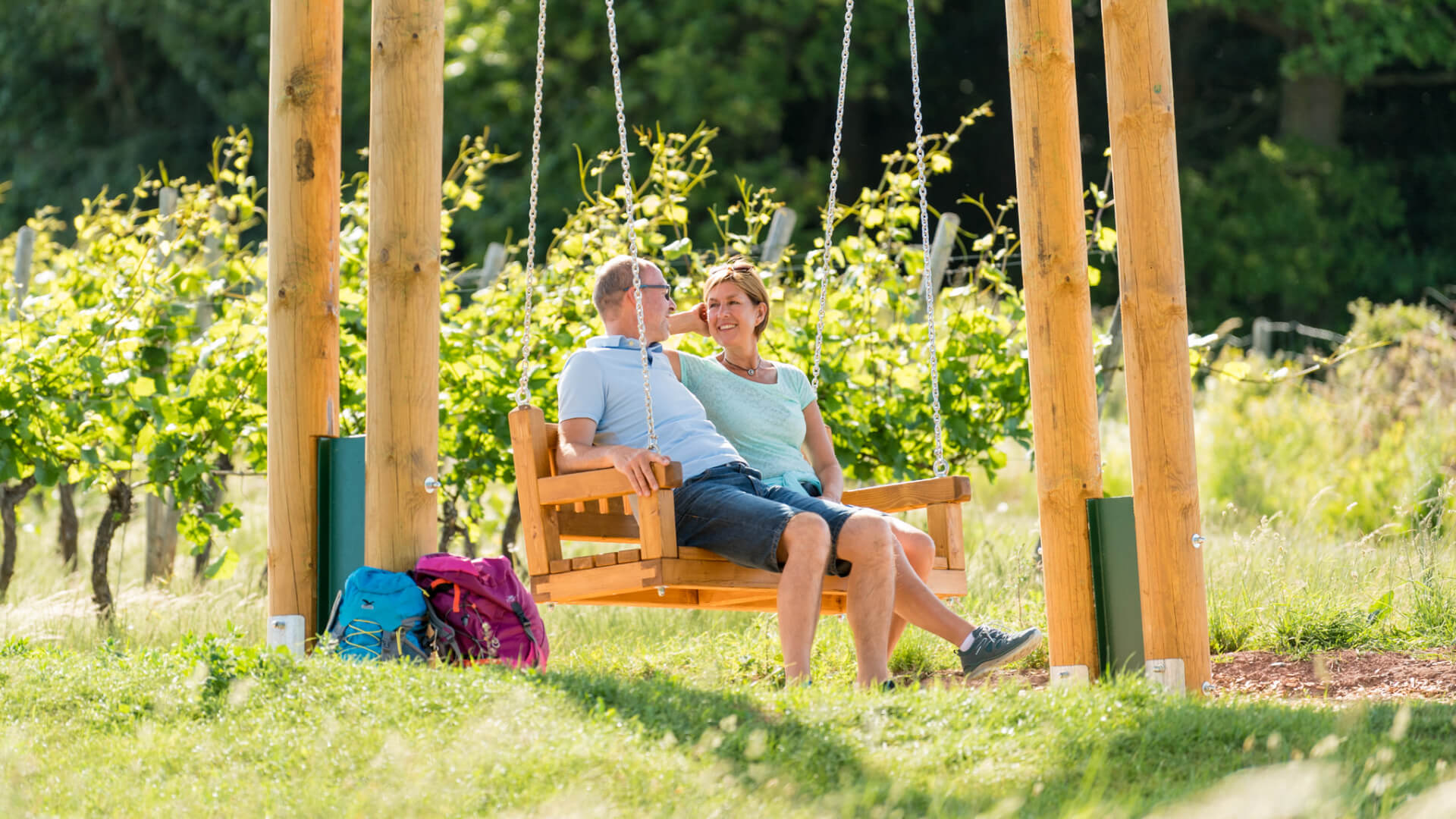 Couple sitting on wooden swing in sunny vineyard with backpacks nearby