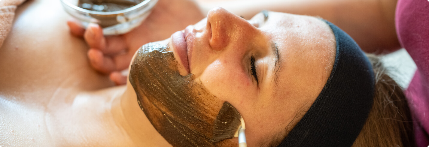 Person receiving facial treatment with dark mud mask at spa