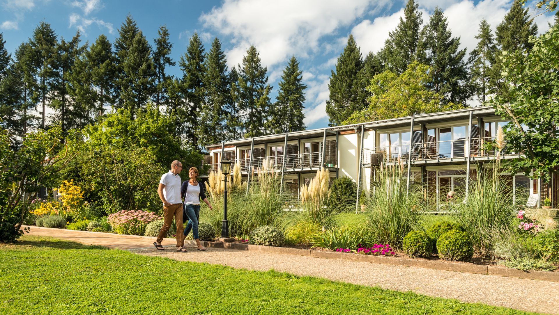 Two people walking near modern house with lush garden and tall trees