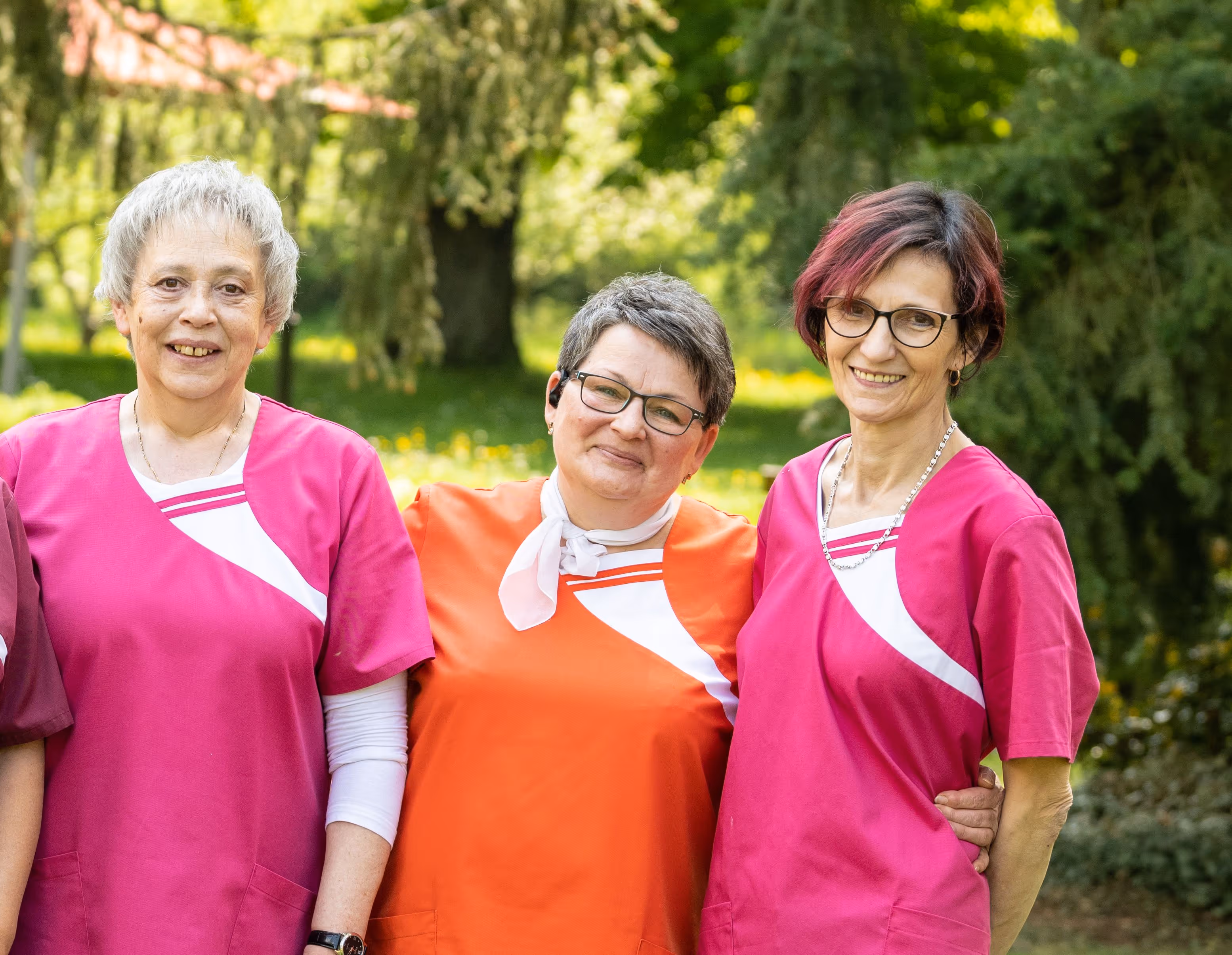 Drei ältere Frauen in rosa und orangefarbenen Hemden lächeln im Freien