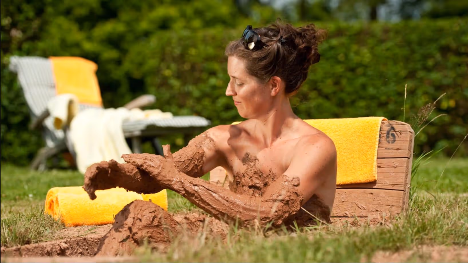 Person relaxing in muddy outdoor bath with yellow towels in garden