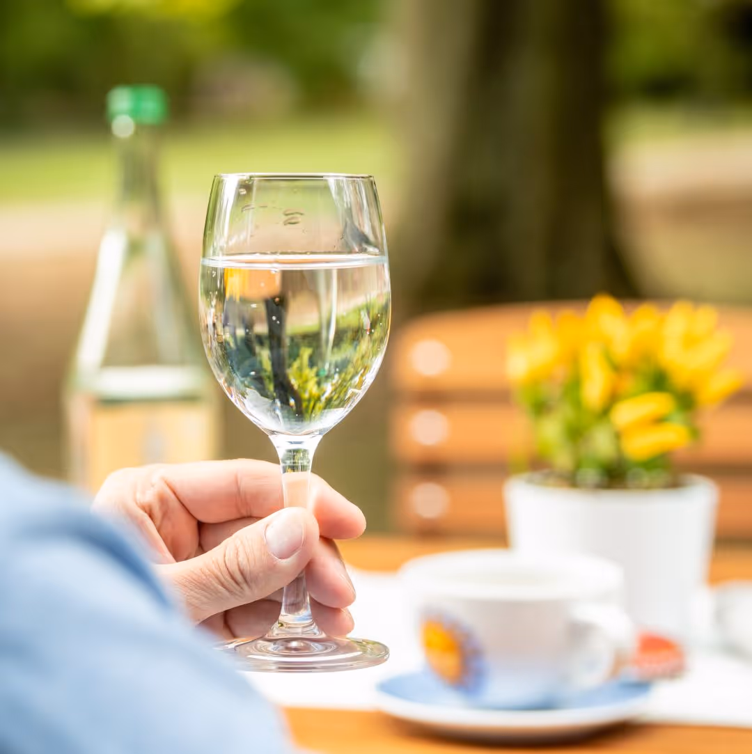 Glas Wasser auf Holztisch mit Blumen und Kaffeetasse im Hintergrund