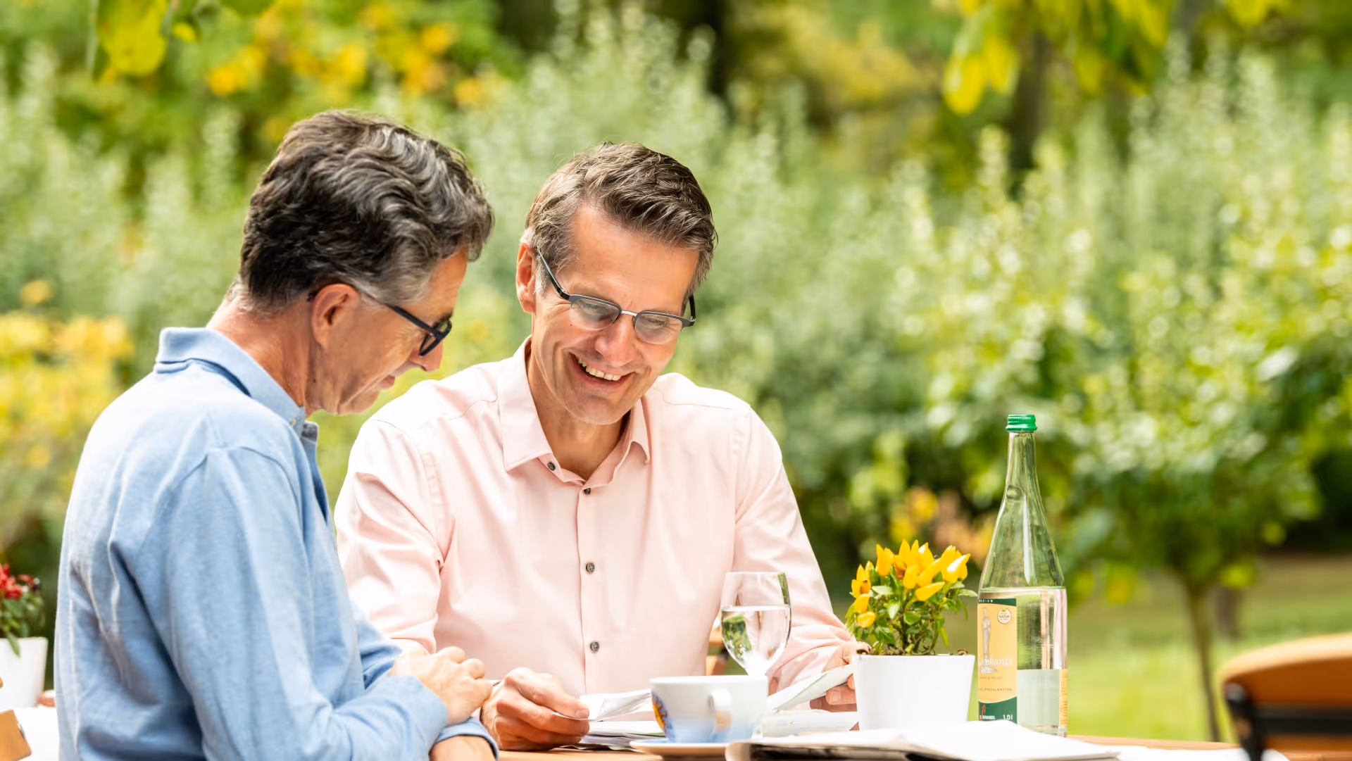 Two middle-aged men laughing together at an outdoor table with flowers