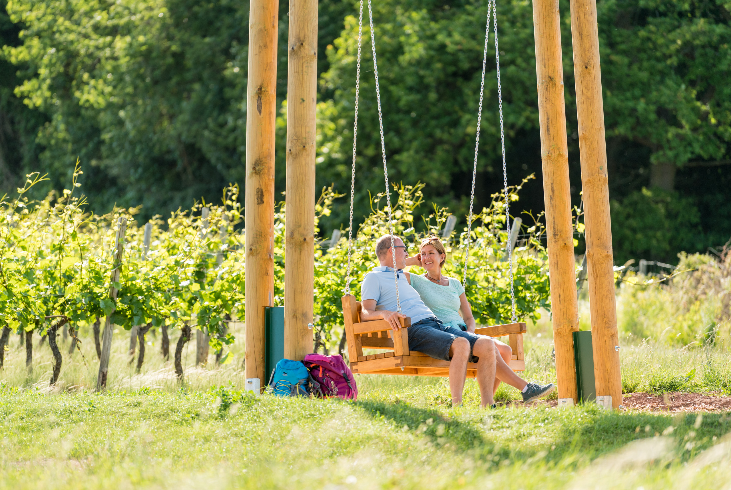 Couple sitting on swing in vineyard with backpacks nearby on sunny day