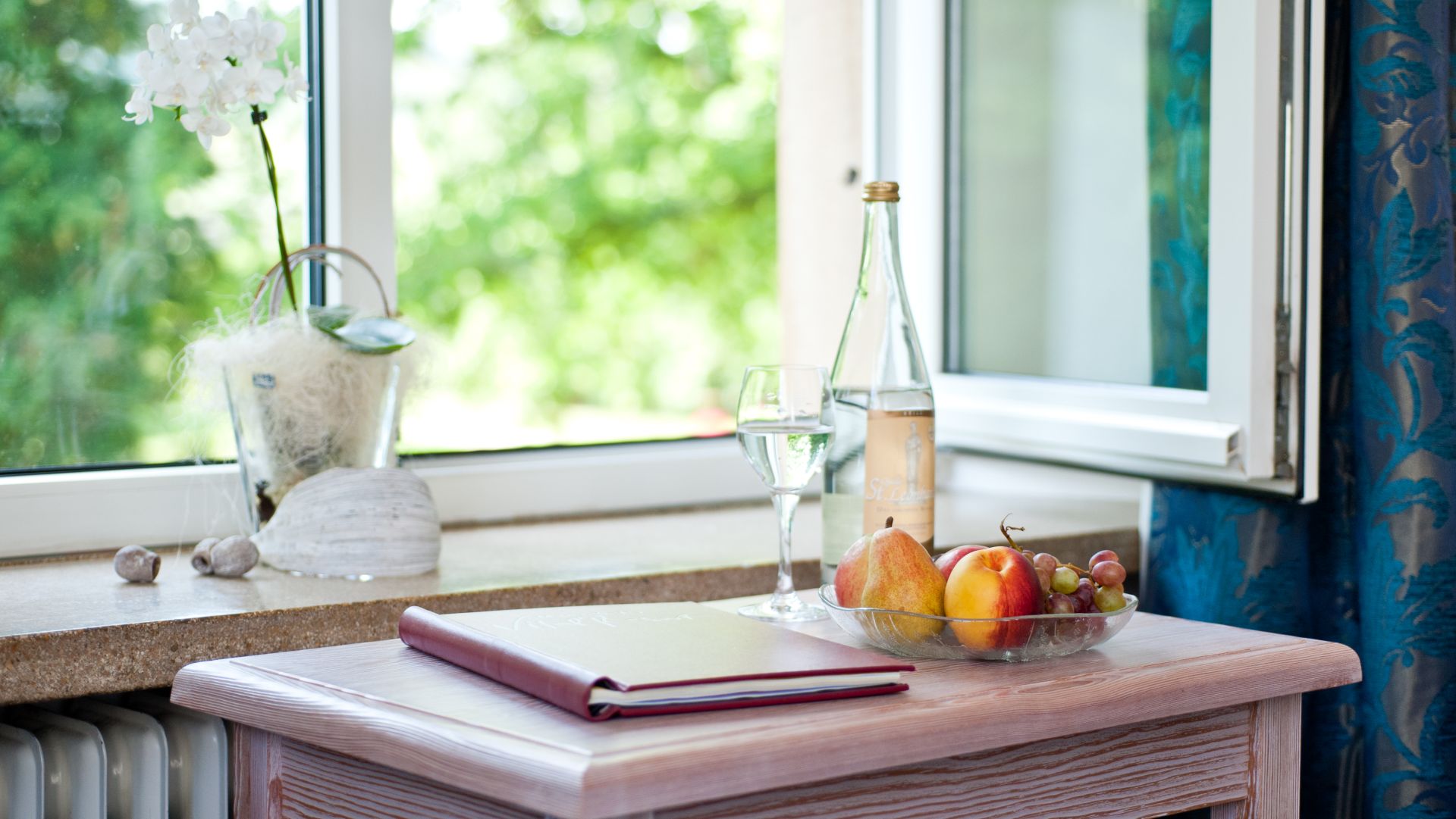 Windowsill with white orchid, fruits, wine glass, and book near open window