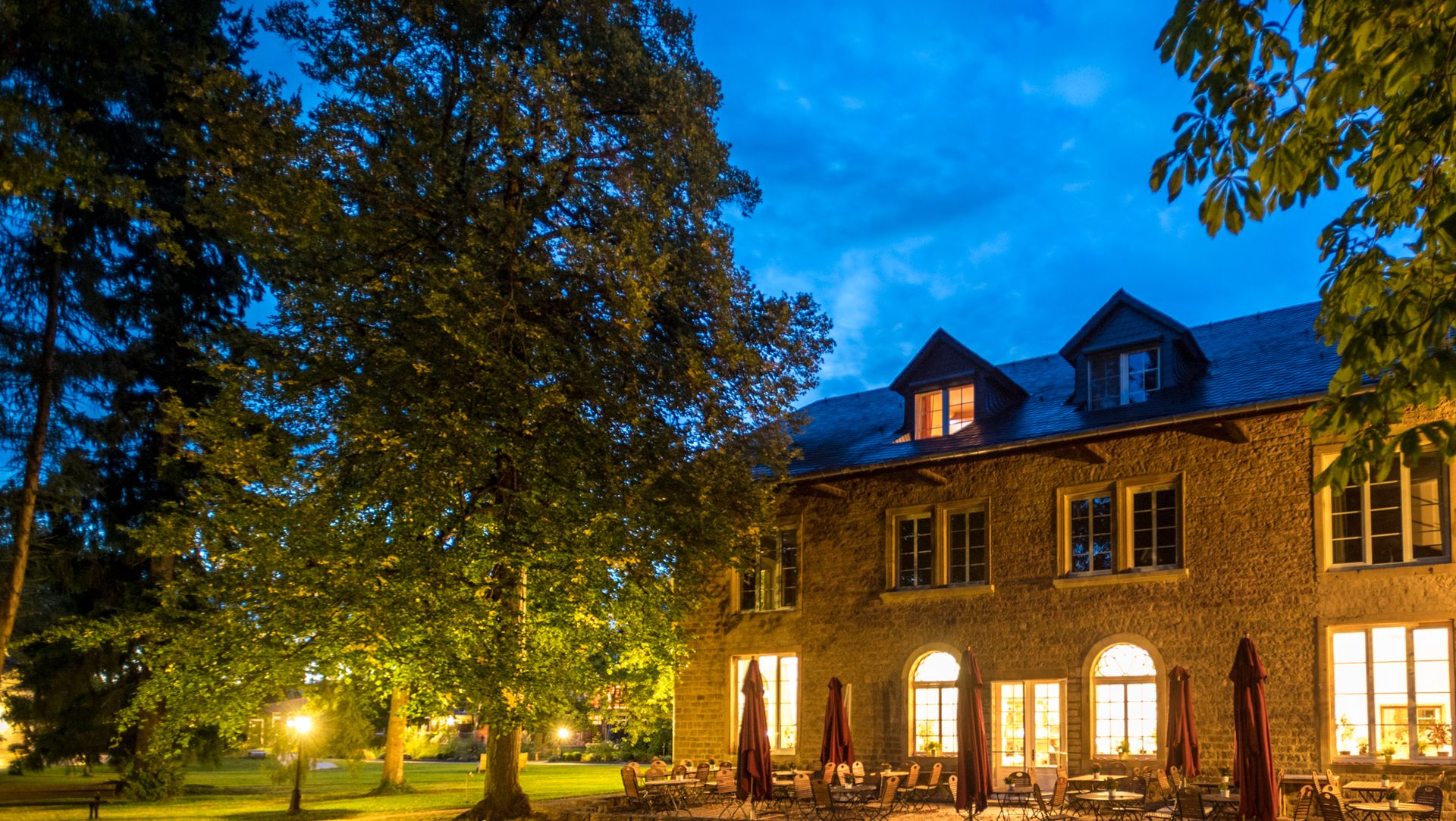 Stone building with warm glowing windows at twilight, surrounded by trees