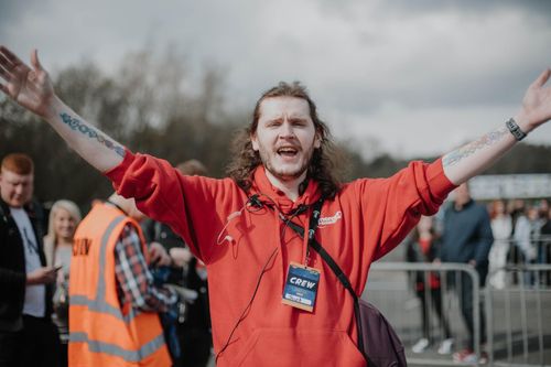 A crew member with arms in the air outside the event wearing a red hoodie