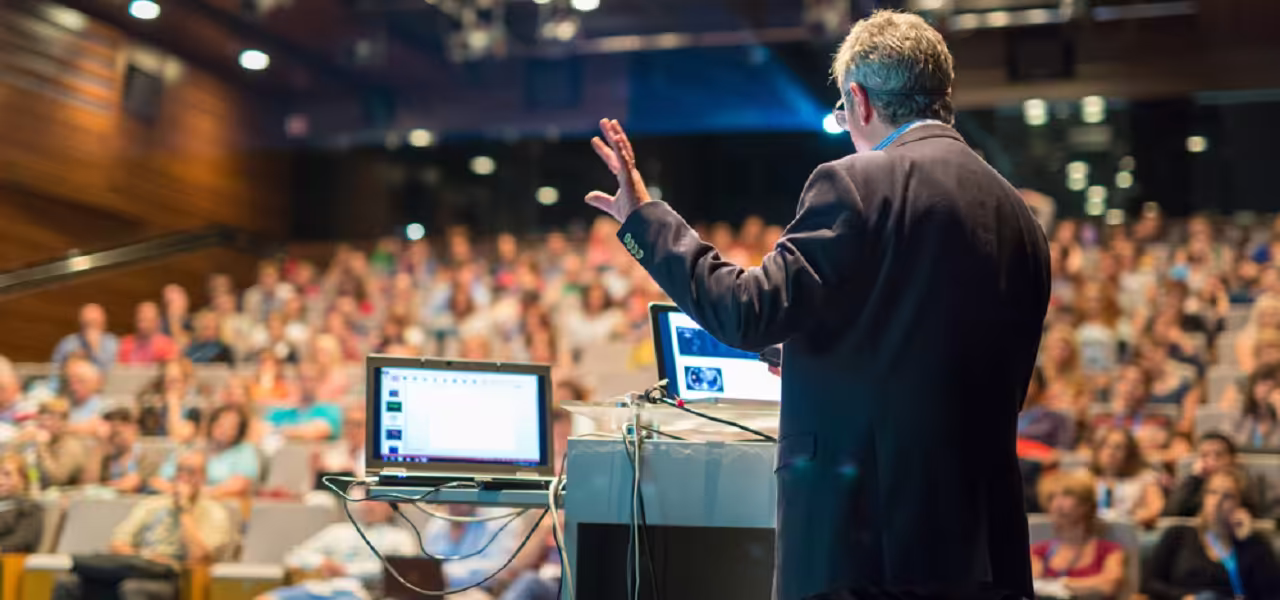 Speaker in front of a packed auditorium presenting with a projector to compare online vs. in-person voting
