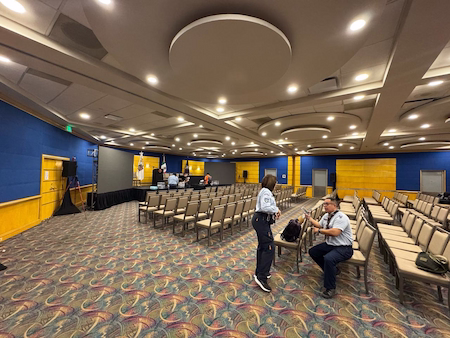 Large conference room with chairs arranged in rows, some people talking near the central aisle and a stage in the background; the place is illuminated and decorated with blue and yellow walls.