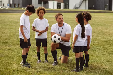 A coach holds a soccer ball and converses with a group of children on a field.