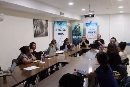 Varias personas sentadas alrededor de una mesa en una sala de reuniones, con portátiles, papeles y botellas de agua, participando en una reunión o taller frente a varias pancartas con textos motivacionales al fondo.