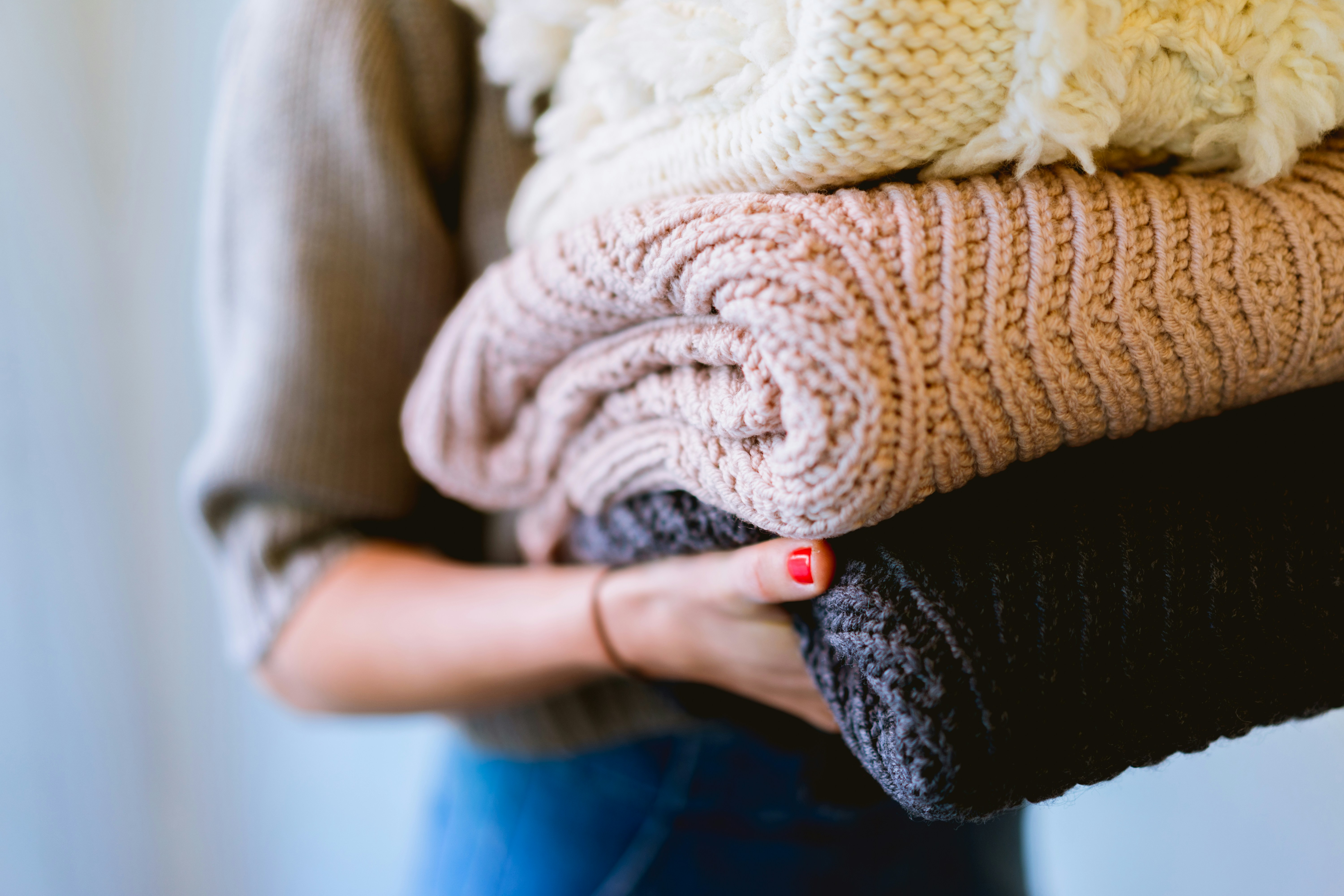 Person holding a stack of folded knitted sweaters in cream, beige, and dark gray colors.