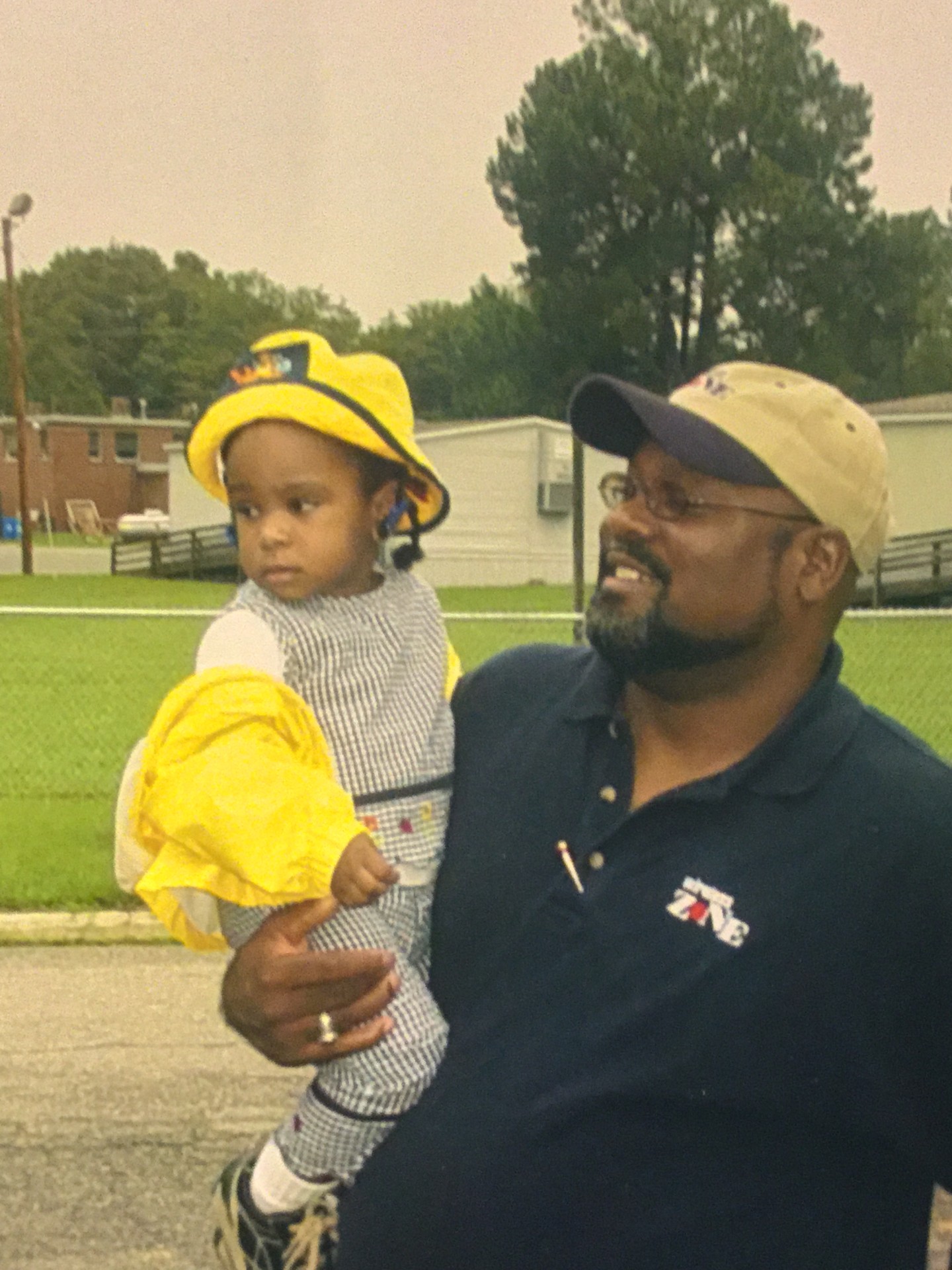 A man in his mid-40s is wearing a navy blue polo shirt and a baseball hat and is smiling holding his young daughter who is a toddler and is wearing a yellow coat with a yellow hat