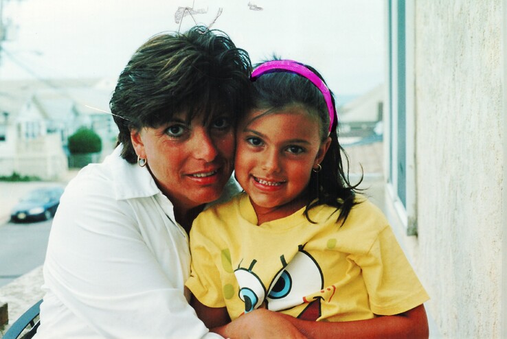 A woman wearing a white blouse hugging her young daughter and smiling at the camera
