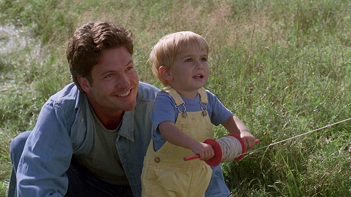 A dad is smiling as he is helping his toddler fly a kite.