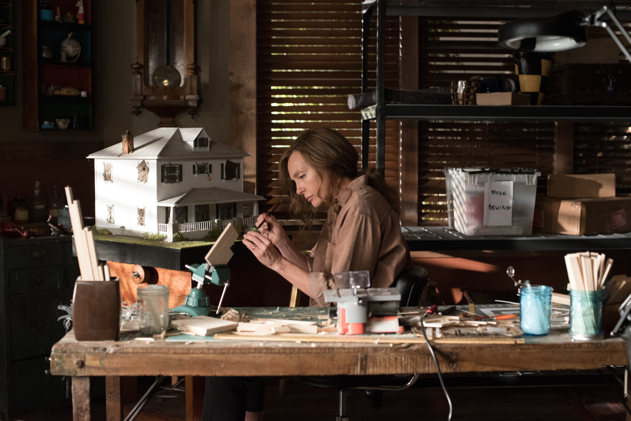 A middle aged woman is sitting at her desk working on a miniature replica of a house.