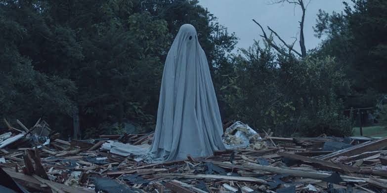 A man covered in a sheet, dressed as a ghost, stands in the wooden rubble of a house that was torn down.