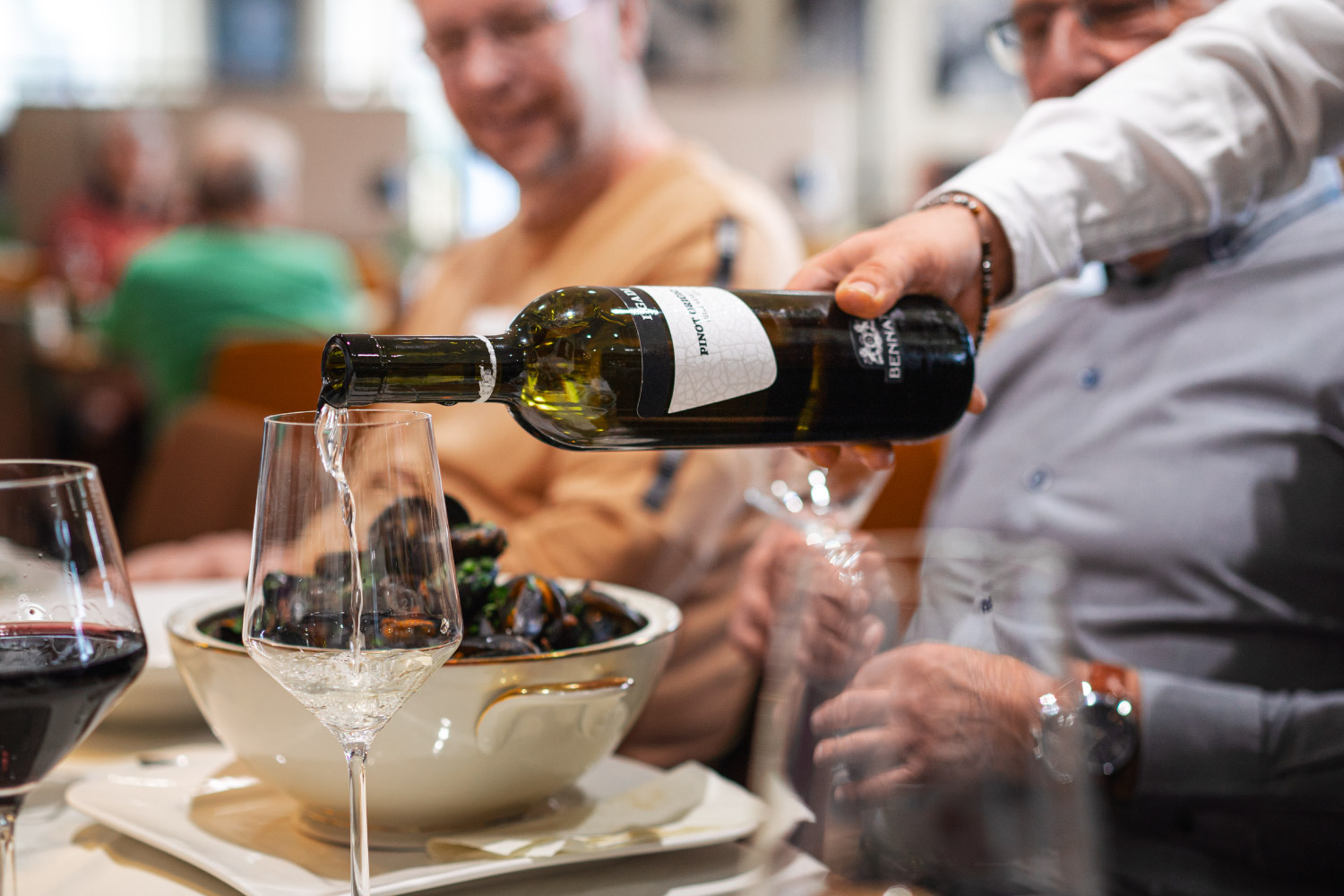 Der Arm einer Person die Weißwein aus einer Flasche beim Essen in ein Weinglas einer Person einschenkt.
