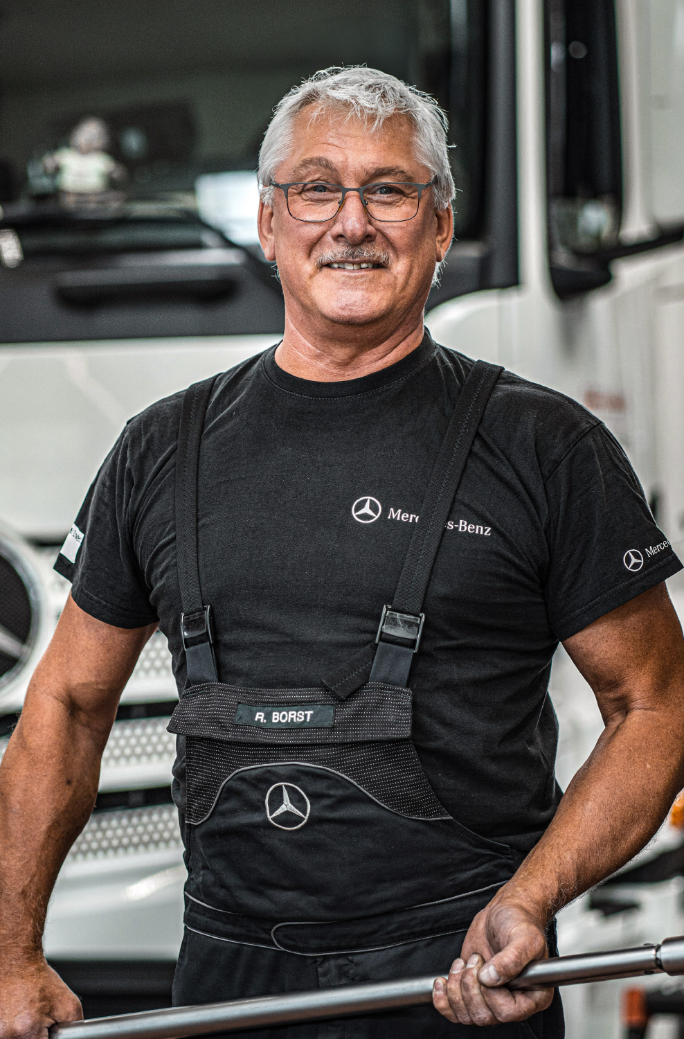 Ein Portrait von einem lächelnden Mann in schwarzer Arbeitskleidung von Mercedes-Benz und einer Brille auf.