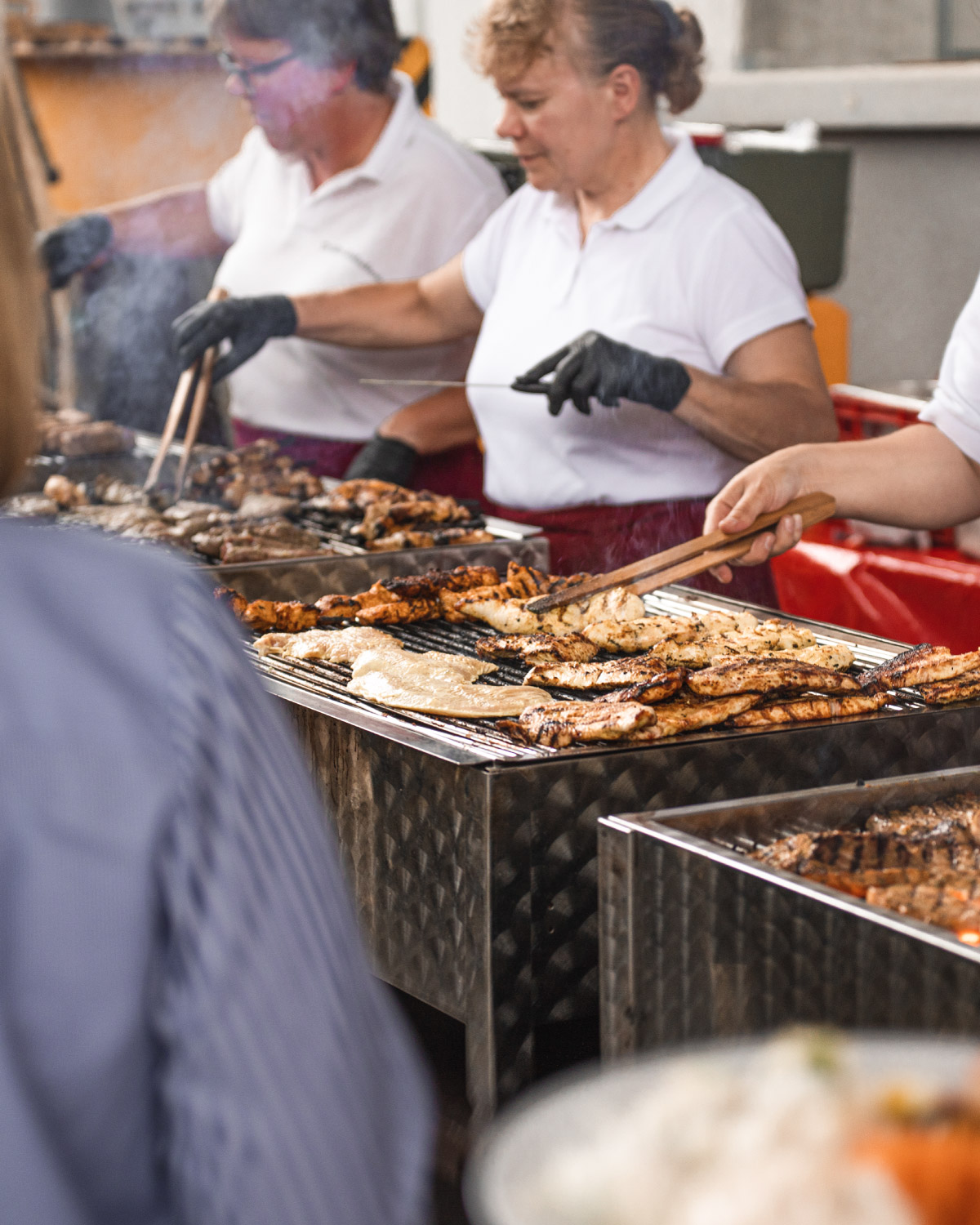 Drei Personen die an einem Grill verschiedene Sorten von Fleisch für eine Firmenfeier grillen.