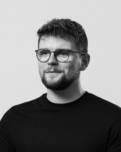Black-and-white portrait of a young man with short curly hair, beard, and glasses wearing a black shirt, looking slightly to the side.