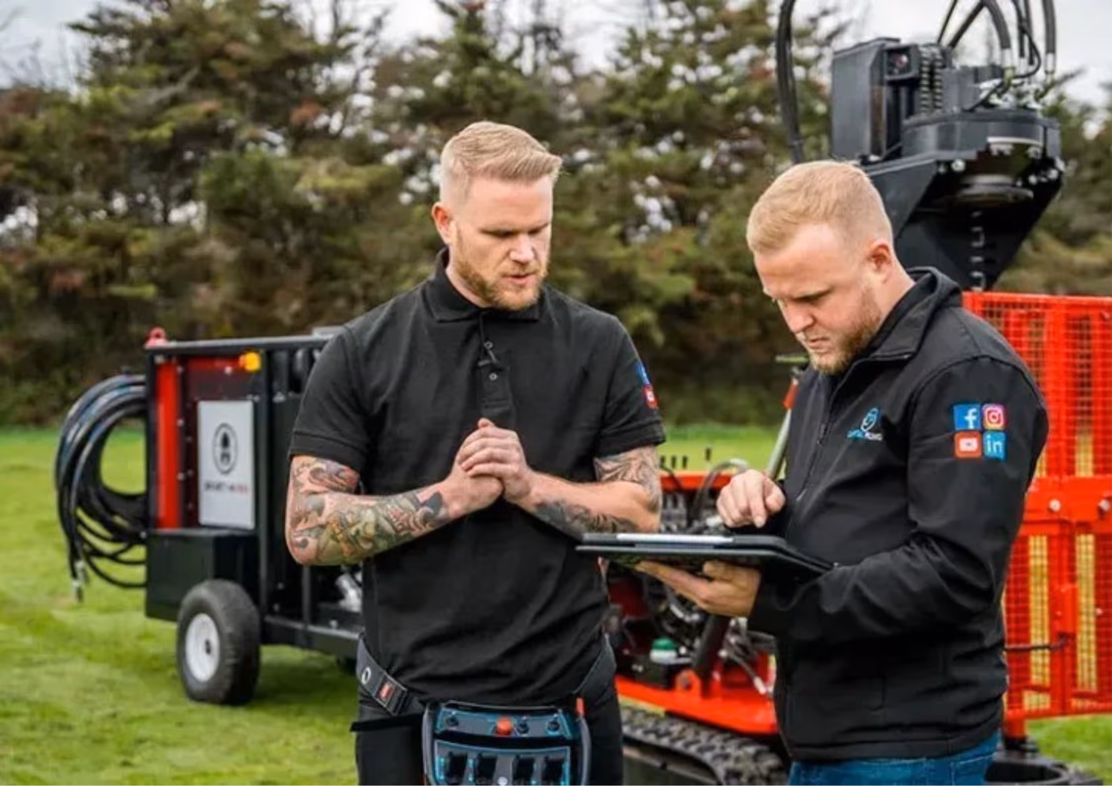 Two men in black workwear operate equipment outdoors; one checks a tablet while the other stands with hands clasped, in front of machinery on grass.