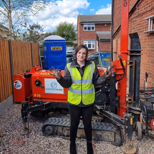 Person wearing a yellow safety vest standing with dirty hands in front of red drilling machinery on gravel between a wooden fence and brick building.