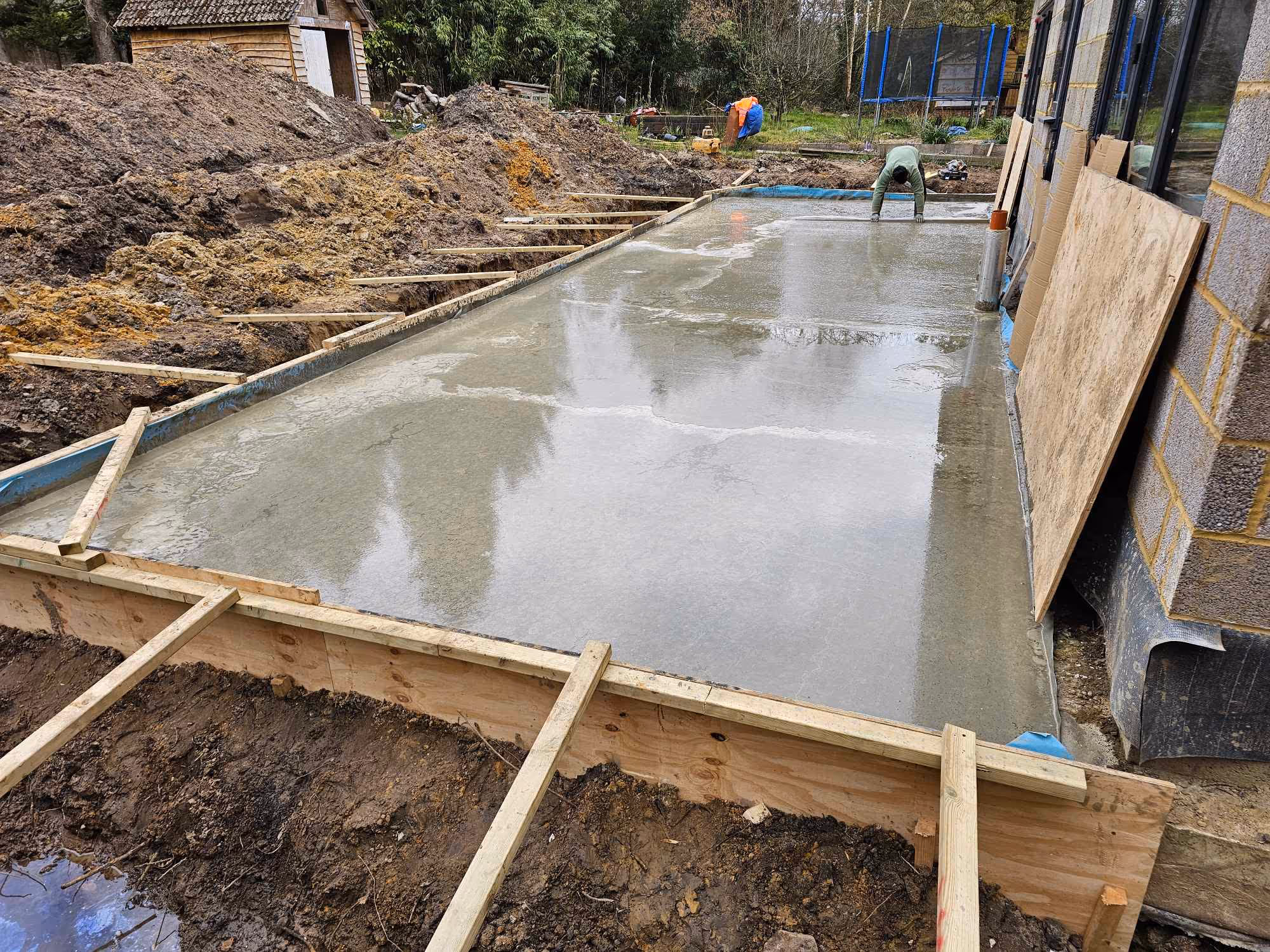 A construction worker smoothing a newly poured concrete slab next to a partially built brick wall with wooden boards for framing.
