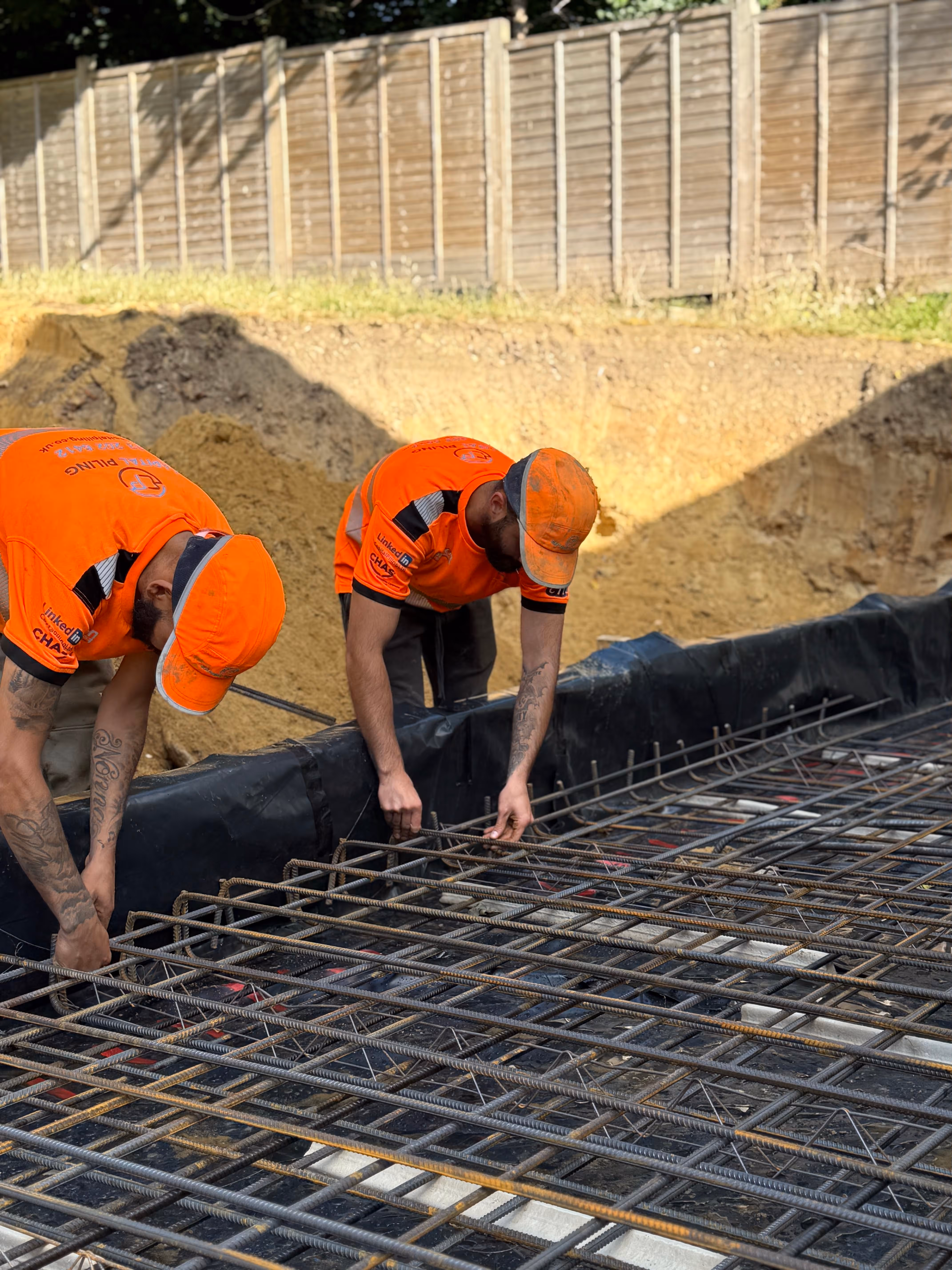 Two construction workers in orange shirts and caps working with steel rebar grid on a foundation site.