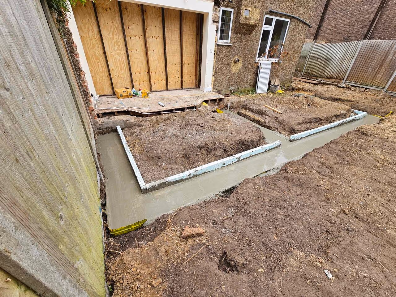 Construction site showing freshly poured concrete footings outlining foundation sections beside a house with boarded-up garage doors.