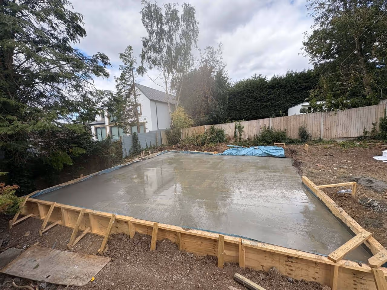 Freshly poured concrete slab foundation with wooden framing around it in a backyard with trees and a house in the background.