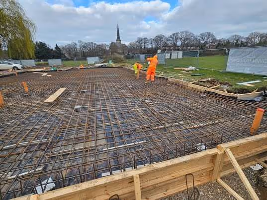 Construction site with metal rebar grid laid out on the ground, wooden framing on the sides, and two workers wearing orange safety clothing.
