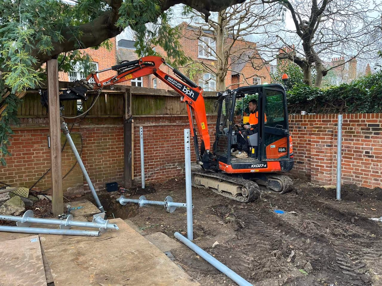 Construction worker operating an orange mini excavator in a backyard with brick walls and metal posts installed in the ground.