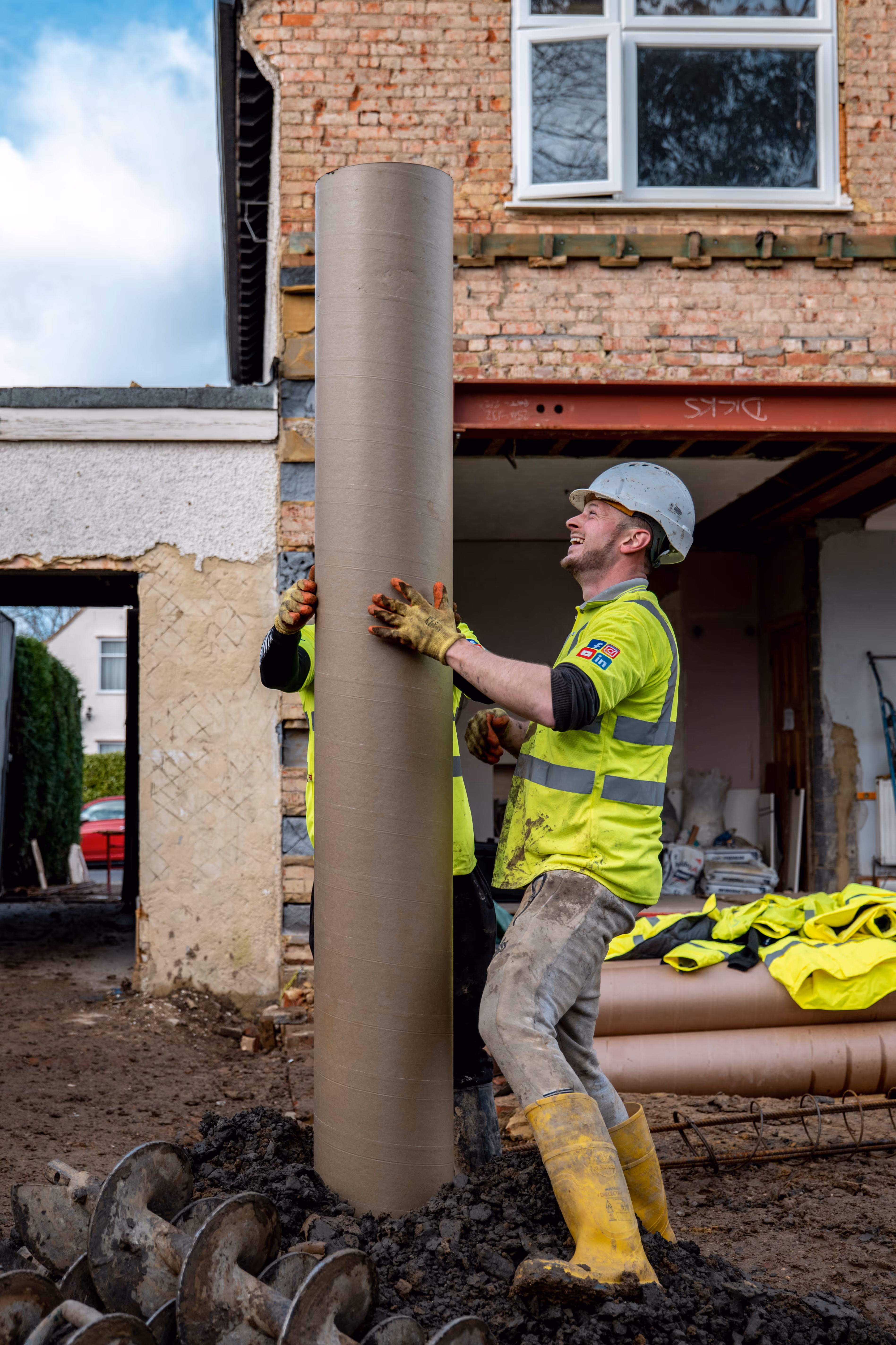 Two construction workers in high-visibility clothing and hard hats handling a large cylindrical pipe on a muddy construction site in front of a partially built brick wall.