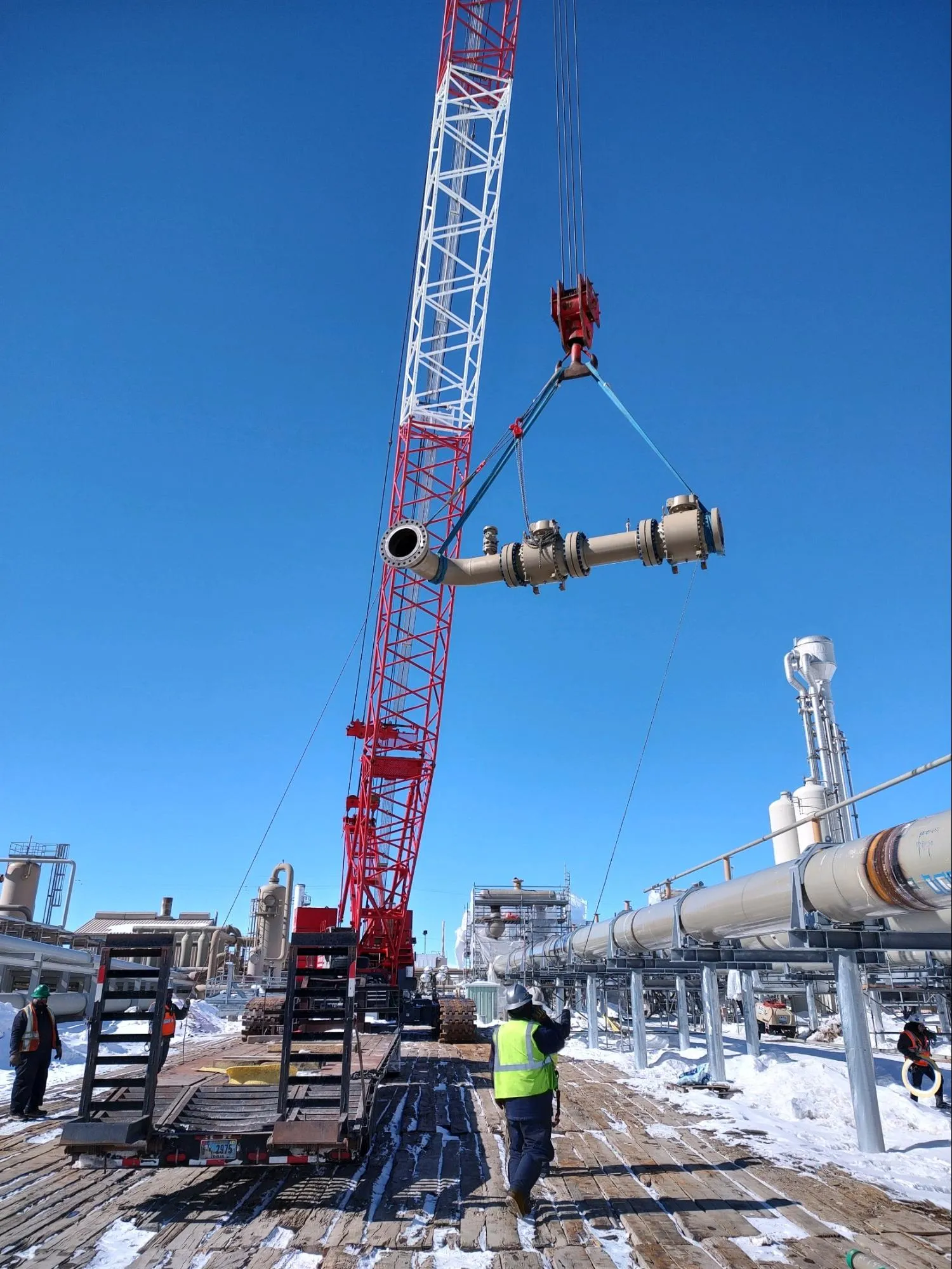 Red and white crane lifting a large industrial pipe at a snowy outdoor construction site with workers wearing safety gear.