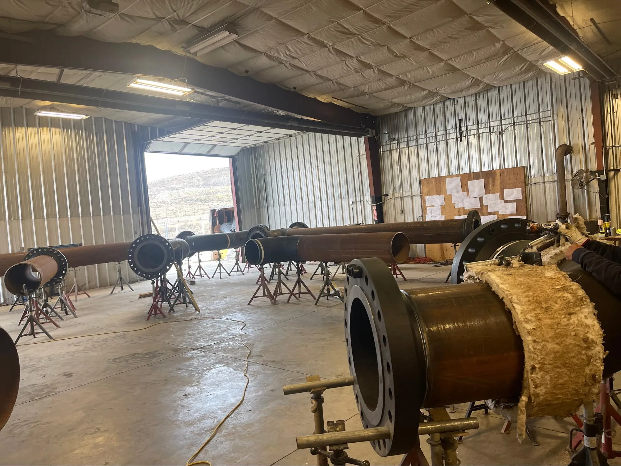 Large metal pipes on stands inside a workshop with a person working on pipe insulation on the right side.