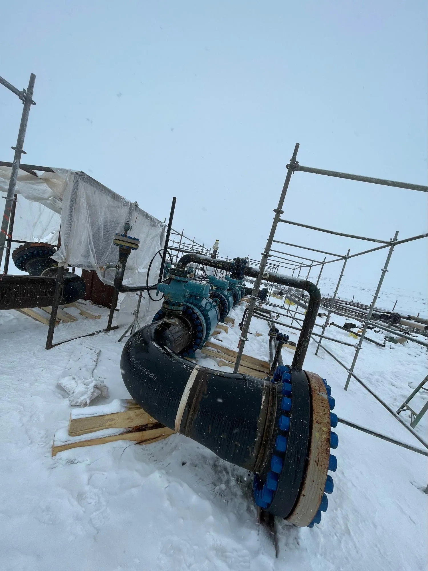 Large industrial pipes with blue valve handles installed outdoors on snow-covered ground surrounded by metal scaffolding.