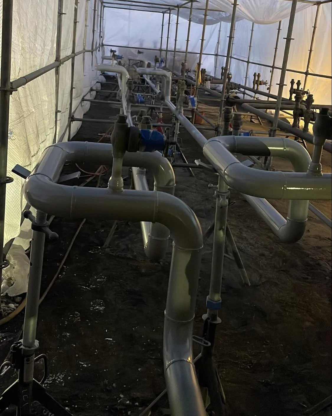 Industrial white piping system with valves under a temporary tent structure on a dirt floor.