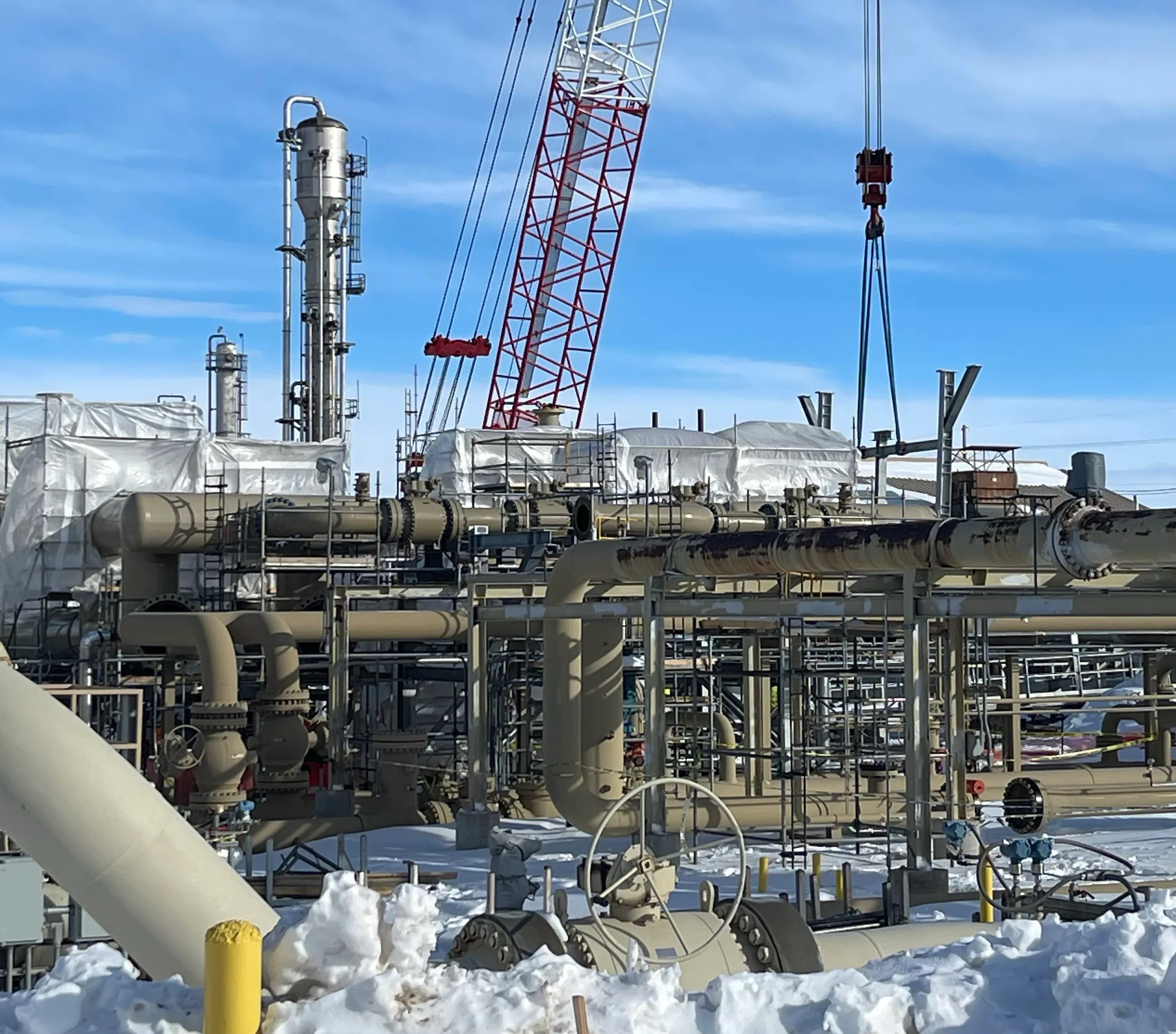 Snow-covered industrial plant with beige pipes, valves, and a red crane against a blue sky.