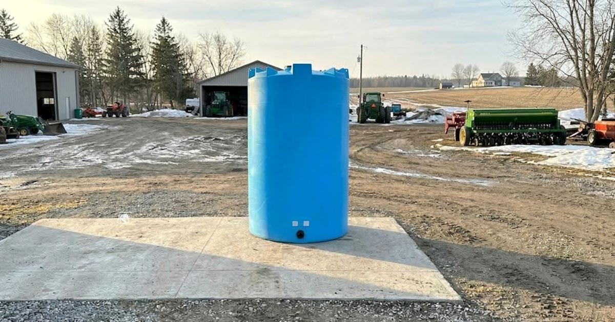 Rotomolded plastic vertical storage tank installed on a concrete pad at a farm, with agricultural equipment and Percheron Plastic branding visible