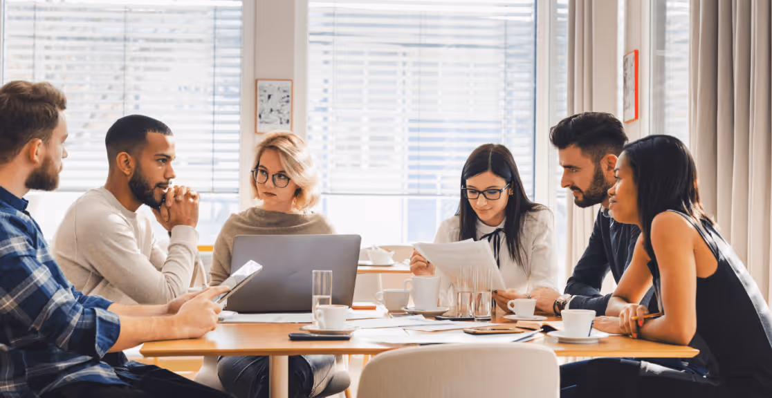 Six diverse young professionals gathered around a table in a bright office, discussing documents and using a laptop.