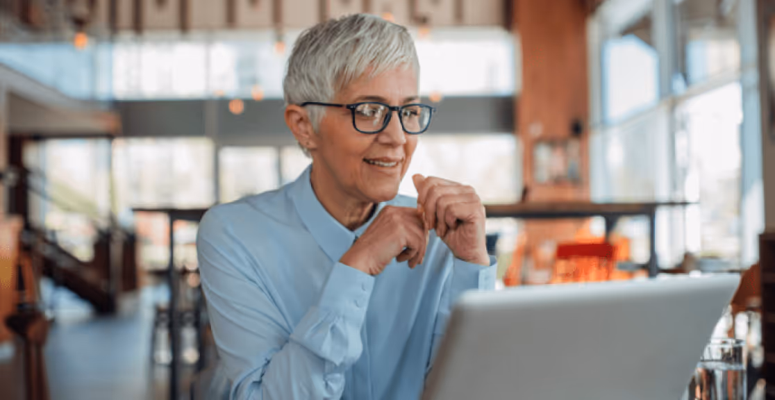 Smiling older woman with short gray hair and glasses sitting in a bright cafe, looking at a laptop screen.