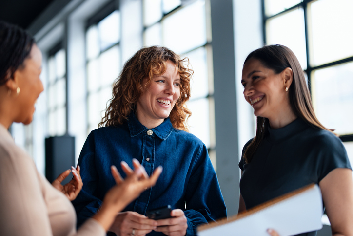 Three diverse women smiling and talking together in a bright office space with large windows, holding a smartphone and documents.