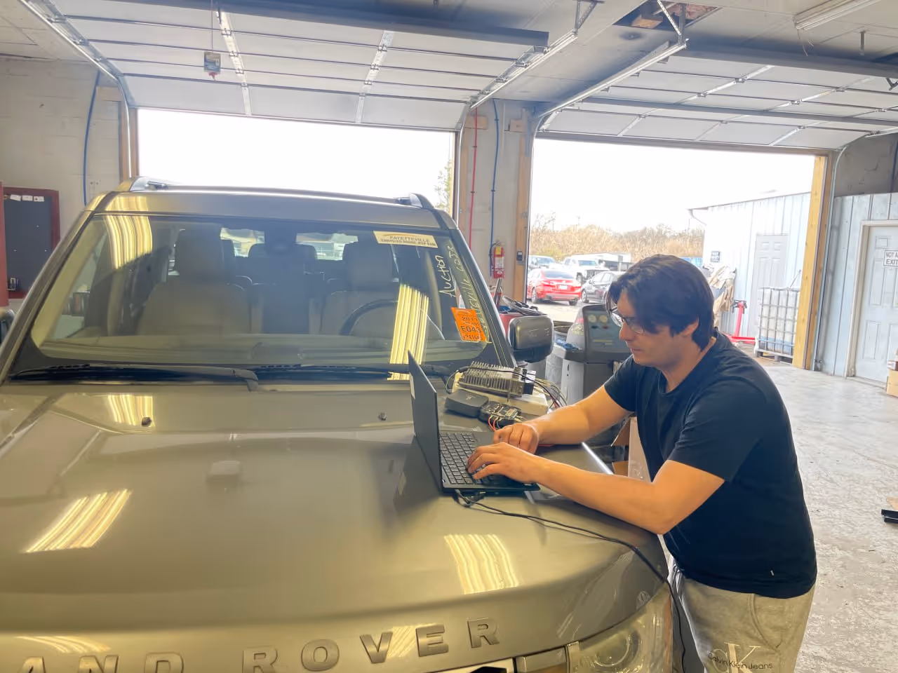 Man working on a laptop placed on the hood of a silver Land Rover inside a garage.