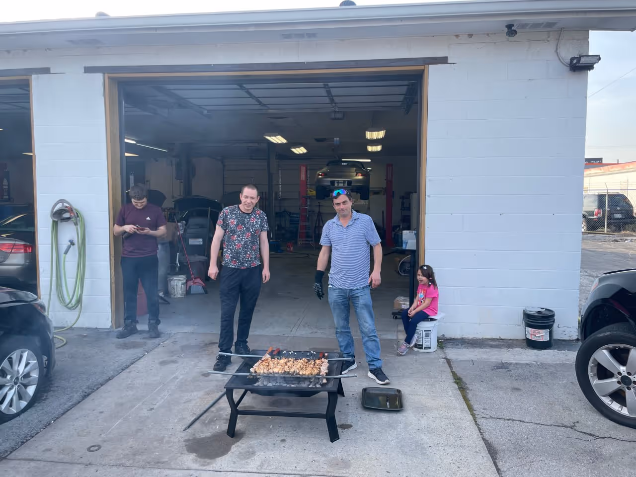 Two men standing and grilling meat skewers outside a garage with an open door, a man looking at his phone inside, and a girl sitting on a bucket.