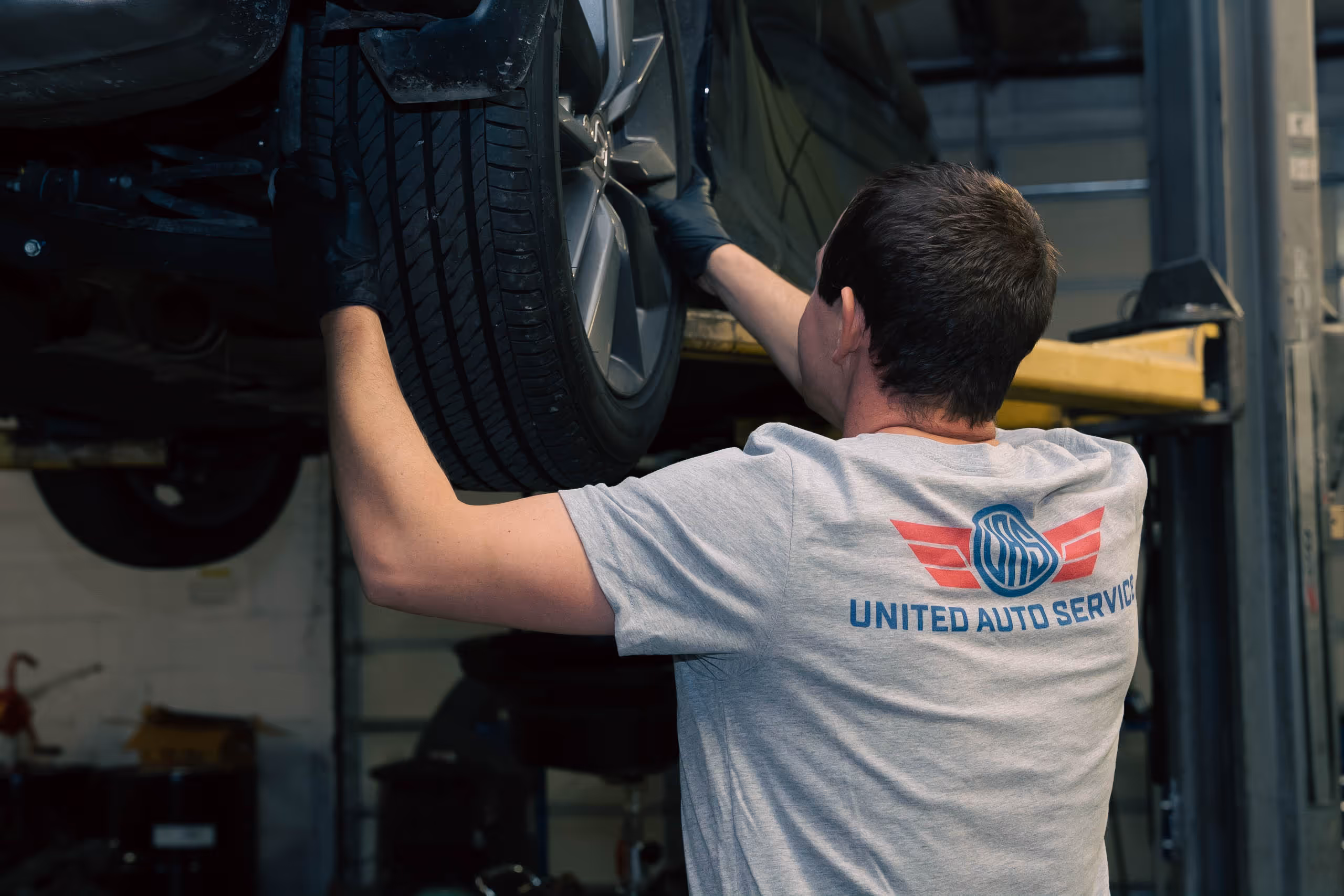 Mechanic wearing a United Auto Service shirt inspecting a car tire lifted on a hoist in a garage.